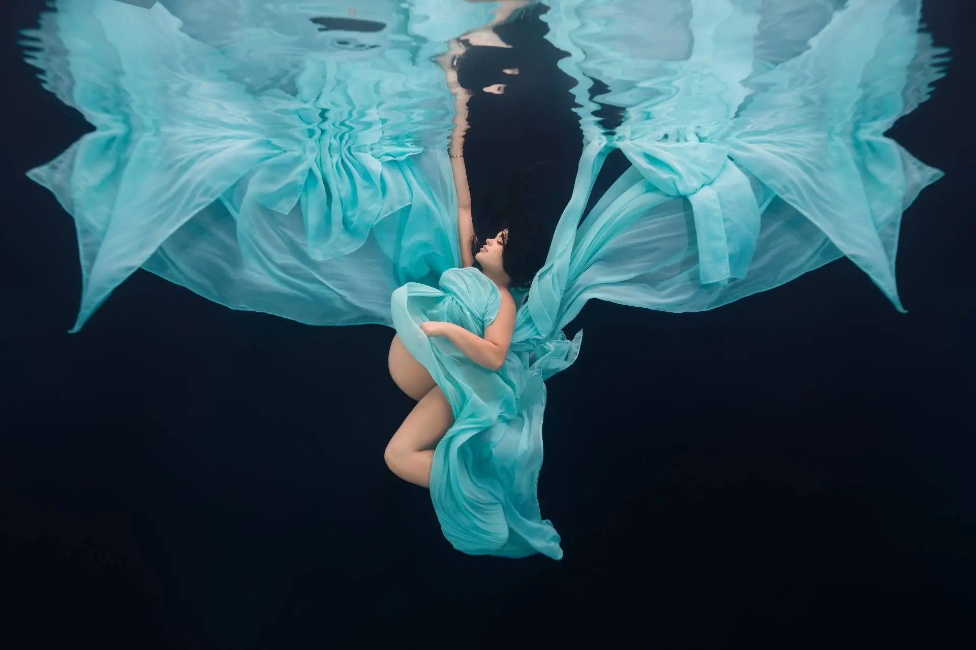 Pregnant woman floats underwater in flowing teal fabric